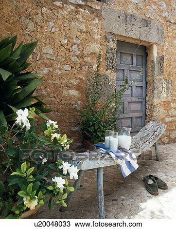 Stock Image - Wooden bench with carafe of milk in front of stone house. Fotosearch