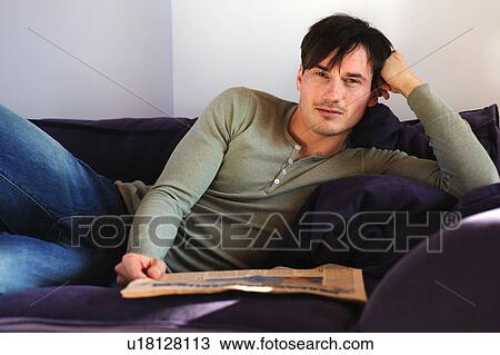Stock Image - Young man on couch reading newspaper. Fotosearch