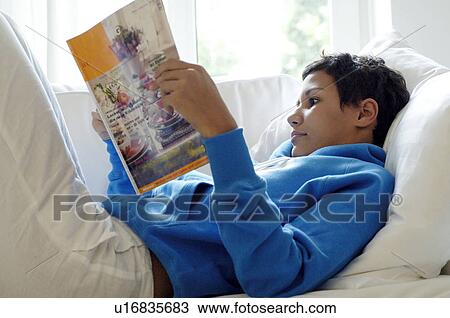 Stock Image - Young woman reading magazine on couch. Fotosearch