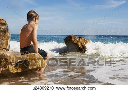 Stock Image - Boy sitting on rock looking out to sea. Fotosearch