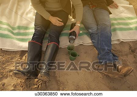 Stock Image - Couple on beach having coffee from flask. Fotosearch
