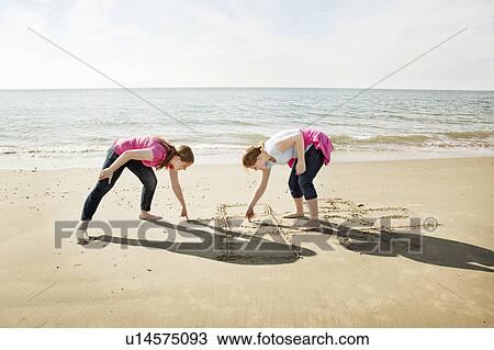 Stock Image - Girls drawing hopscotch in sand on beach. Fotosearch