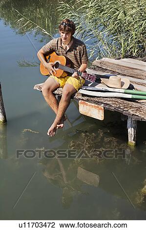 Stock Image - Man sitting on jetty playing a guitar. Fotosearch