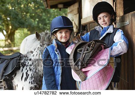 Stock Image - Two girls holding saddles with pony. Fotosearch