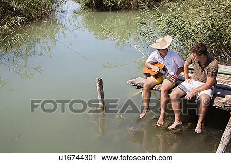 Stock Image - Two men sit chatting on lake jetty. Fotosearch