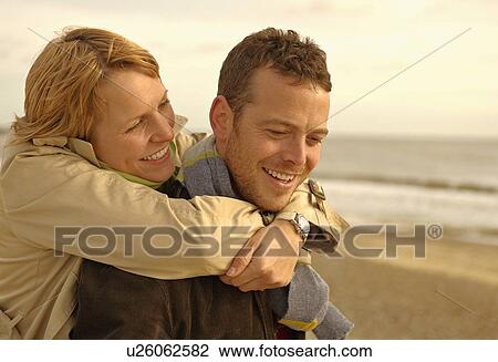 Stock Image - Woman embracing man from behind on beach. Fotosearch