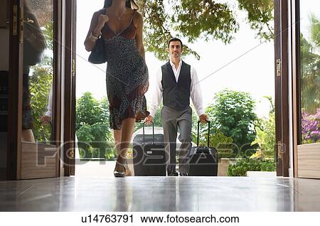 Stock Image - Woman entering hotel man carrying bags. Fotosearch