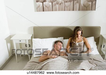 Stock Image - Woman using laptop in bed, man asleep. Fotosearch
