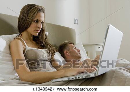 Woman using laptop in bed, man asleep View Large Photo Image Stock Photo - Woman using laptop in bed, man asleep. Fotosearch