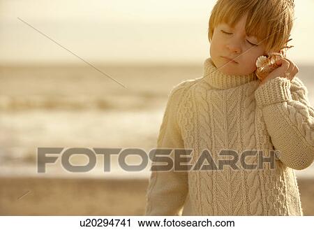 Stock Image - Young boy by sea, listening in seashell. Fotosearch