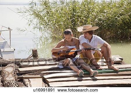 Stock Image - Young couple sitting together on jetty. Fotosearch