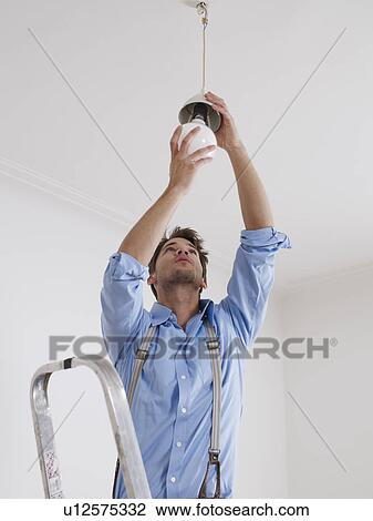 Stock Image - Young man on ladder screws in light bulb. Fotosearch
