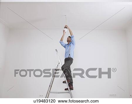 Stock Image - Young man on ladder screws in light bulb. Fotosearch