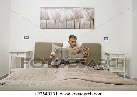 Stock Image - Young man reading newspaper in bed. Fotosearch
