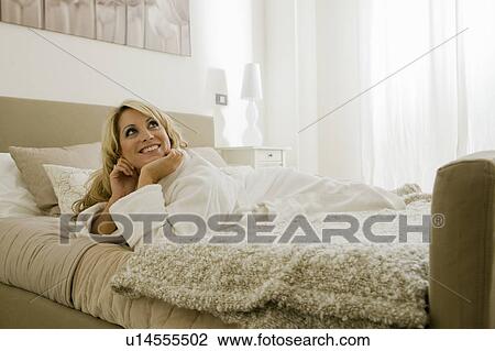 Stock Image - Young woman in bathrobe lying on bed. Fotosearch