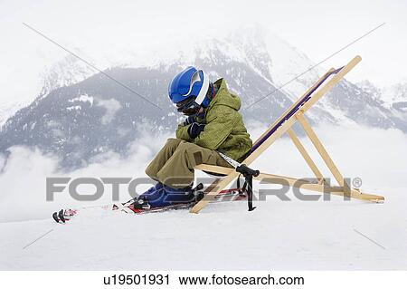 Stock Image - Child with skis sitting on deck chair. Fotosearch