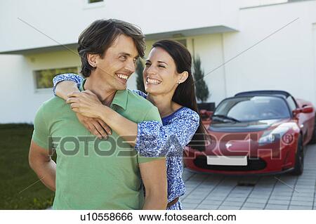 Stock Photograph - Couple in front of house + electric car. Fotosearch