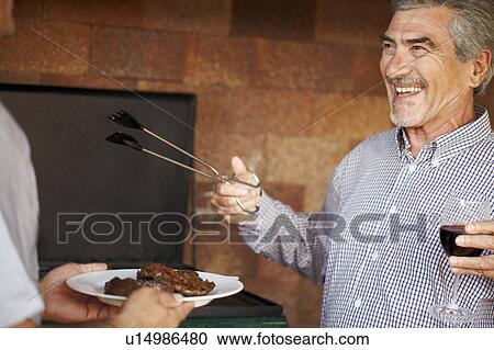 Stock Image - Happy older man serving up meat from BBQ. Fotosearch