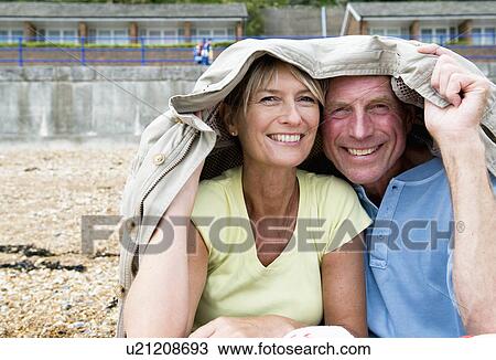 Stock Image - Seniors shelter under coat on beach. Fotosearch