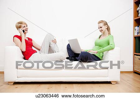 Stock Photograph - Two women using laptop and phone on sofa. Fotosearch