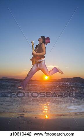 Stock Image - Young man jumps with guitar at sunset. Fotosearch