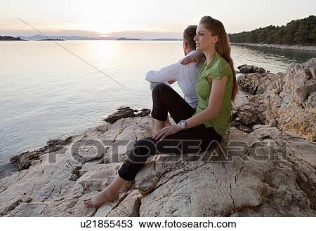 Couple watching sun set at rocky beach View Large Photo Image Stock Image - Couple watching sun set at rocky beach. Fotosearch