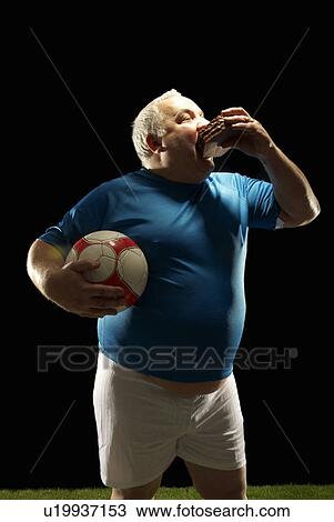 Stock Image - Large footballer holding ball and eating. Fotosearch