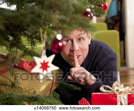 Stock Image - man hiding presents under christmas tree. Fotosearch