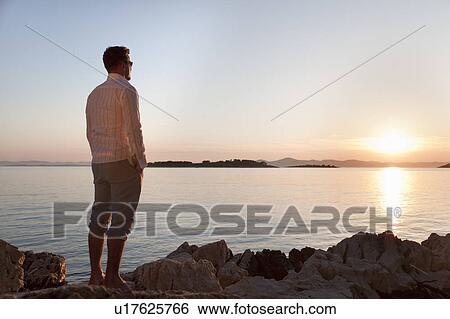 Man watching sun set at rocky beach View Large Photo Image Stock Photograph - Man watching sun set at rocky beach. Fotosearch