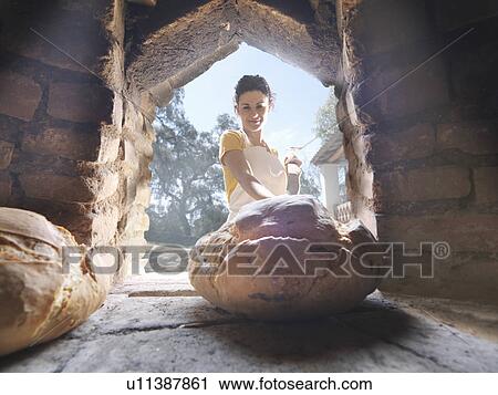 Stock Image - Woman taking bread out of wood oven. Fotosearch
