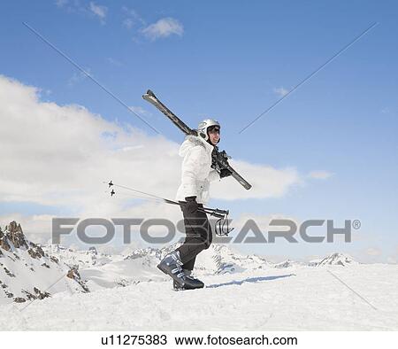 Stock Image - Woman walking up hill in snow, with skis. Fotosearch