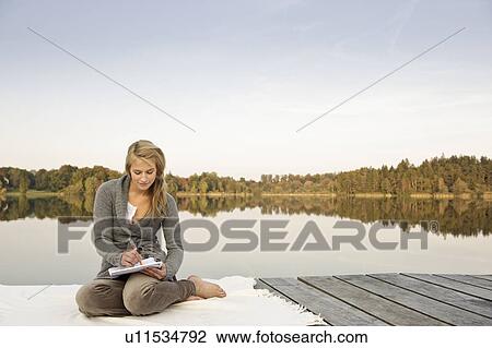 Stock Image - Young woman writing, sketching, by lake. Fotosearch