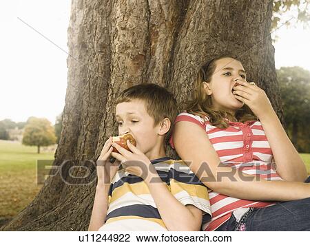 Boy and girl eating apples against tree View Large Photo Image Stock Image - Boy and girl eating apples against tree. Fotosearch