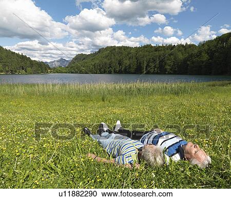 Stock Image - Senior couple lying in field by lake. Fotosearch