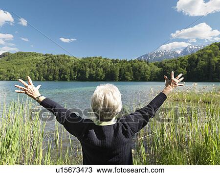 Stock Image - Senior woman standing by lake, rear view. Fotosearch