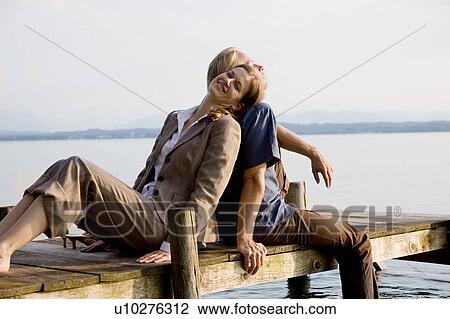Stock Image - woman and man sitting on pier at lake. Fotosearch