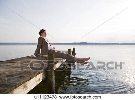 Stock Photo - woman sitting on pier at lake. Fotosearch