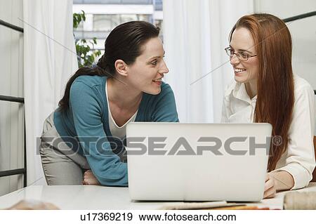 Stock Photo - 2 women smiling at each other, behind a laptop. Fotosearch