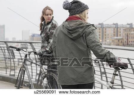 Stock Image - 2 young women pushing bikes. Fotosearch