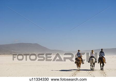 Stock Image - 3 people riding horses on the beach. Fotosearch