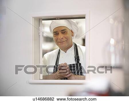 Stock Image - Chef looking through hatch in restaurant. Fotosearch