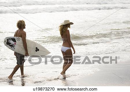 Stock Image - Guy and girl wlking on beach in sunset, him with surf board. Fotosearch