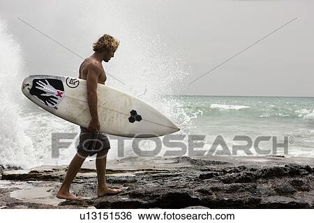 Guy with surfboard walking on rocks. Ocean behind him and water splashing View Large Photo Image Stock Photograph - Guy with surfboard walking on rocks. Ocean behind him and water splashing. Fotosearch