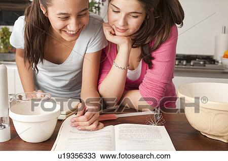 Stock Image - teenage girls looking at recipe in kitchen. Fotosearch