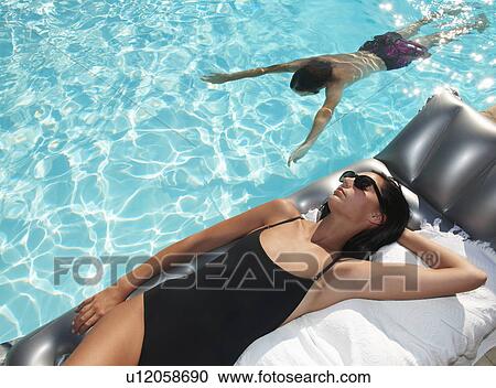 Stock Image - Woman tanning by the pool, man swimming. Fotosearch