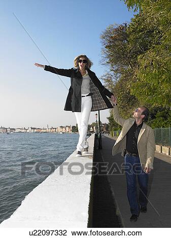 Stock Image - Woman walking on a low wall, by the sea. Fotosearch