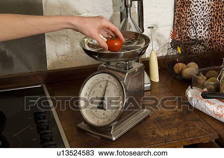 Stock Image - Woman weighing tomato in kitchen. Fotosearch