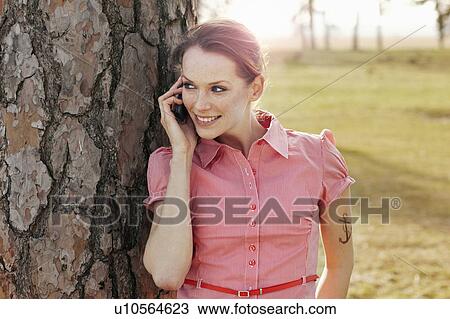 Stock Image - Women with Cellphone leaning on tree. Fotosearch