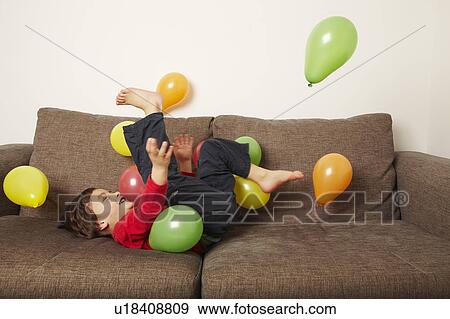 Stock Photo - Young boy lying on sofa kicking balloons. Fotosearch