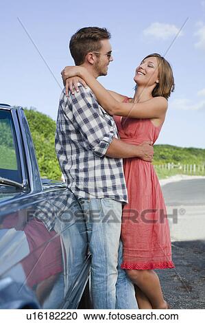 Stock Image - Couple hugging by convertible. Fotosearch